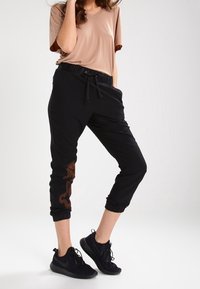 Black jogger pants with elastic cuffs, featuring a drawstring waist and a dragon pattern in brown on the left leg. Paired with a beige top.