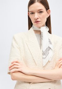 Femme aux cheveux bruns lisses portant un blazer crème texturé et une écharpe blanche à motifs nouée autour du cou, les bras croisés.