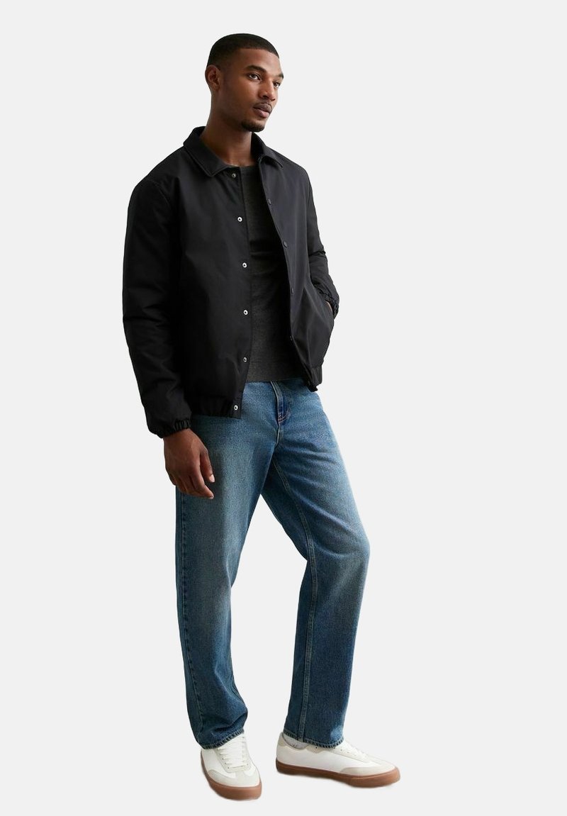 Giacca bomber nera con bottoni a pressione, indossata sopra una maglietta scura. Abbinata a jeans con gamba dritta blu chiaro e sneakers bianche con dettagli beige.