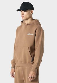 Brun hættebluse med kengurulomme, ribbede manchetter og logodetaljer. Båret med matchende bukser; modellen har en sort cap på.