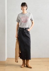 Graues T-Shirt mit rotem Druck, schwarze Denim-Maxiröhre und hellbraune High Heels. Hält eine hellbraune Jacke und steht auf einem Holzfußboden.