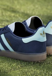 Σκουρομπλε αθλητικά παπούτσια Adidas Gazelle με ανοιχτό μπλε ρίγες και ταμπέλα στη φτέρνα, τοποθετημένα σε πράσινο γρασίδι με υφασμένες λαστιχένιες σόλες.