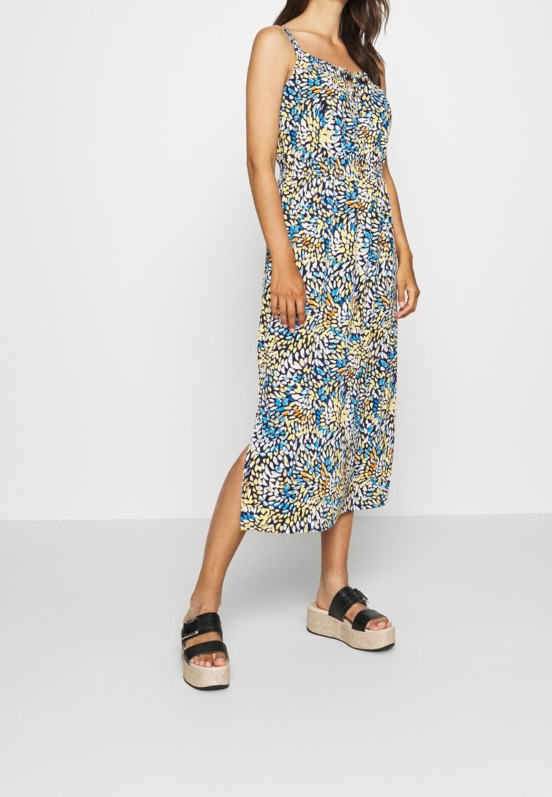 Femme portant une robe midi sans manches à motifs floraux multicolores bleu, jaune et blanc, avec des sandales compensées noires.