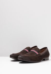 Mocassins en cuir suedé marron avec un talon bas, présentant un design à bandes avec des rayures rouges, blanches et bleues sur le dessus. Intérieur en cuir lisse.