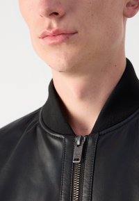 Zwarte leren bomberjack met een zwarte geribde kraag en een zilveren ritsdetail, met een gladde textuur en minimale stiksels.