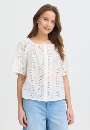 Jeune femme aux longs cheveux bruns portant un chemisier blanc à manches courtes en broderie anglaise et un jean bleu clair, souriant doucement.