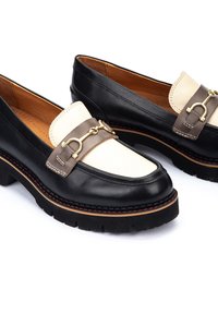 Mocasines de cuero negro y crema con una plataforma baja, que presentan un acento de hardware dorado y detalles de costura en contraste.