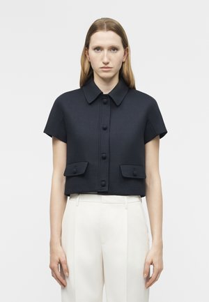 Ženska, oblečena v kratkoročno črno jakno z gumbi in dvema sprednjima žepoma ter bele hlače z visokim pasom, stoji na enostavni podlagi.