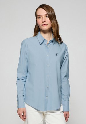 Camiseta de manga larga azul claro, hecha de algodón, con un cuello abotonado y un logo bordado en el lado izquierdo del pecho. Textura suave.