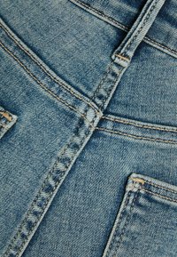 Primer plano de tela vaquera que muestra un color azul claro con costuras amarillas contrastantes, costuras visibles de bolsillo y un acabado texturizado.