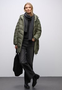 Abrigo acolchado verde con capucha, bufanda gris oscuro, suéter negro y jeans de pierna ancha. Botines negros y un bolso negro completan el conjunto.