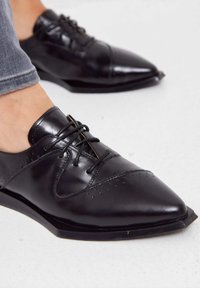 Chaussures habillées en cuir noir avec un bout pointu, dotées de détails perforés, d'un laçage et d'une texture lisse. Semelle plate.