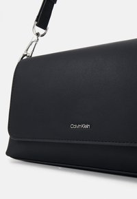 Bolso de mano negro de cuero sintético con forma estructurada, herrajes plateados y logo "Calvin Klein" en relieve. Incluye correa ajustable.