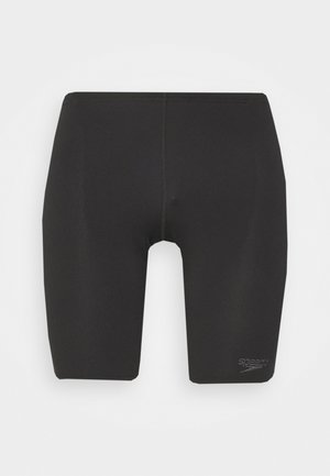 Shorts de bain noirs coupés au genou, fabriqués dans un matériau extensible, avec une texture lisse et des coutures minimales, arborant un petit logo sur la jambe gauche.