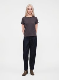 Jeune femme aux longs cheveux blonds, debout face à nous, portant un t-shirt gris foncé, un jean large foncé et des bottines marron.