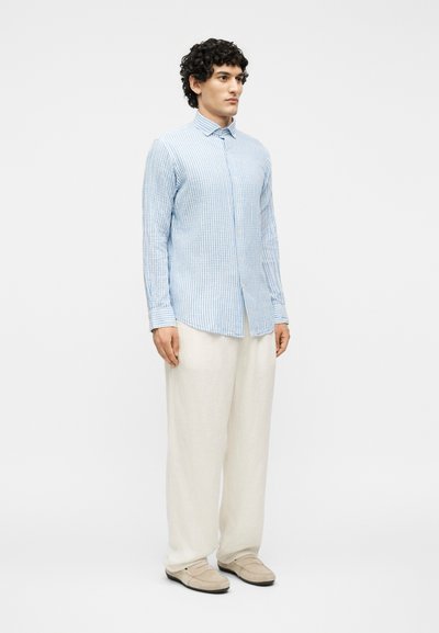 Άνδρας στέκεται, φοράει γαλάζιο ριγωτό πουκάμισο με κουμπιά, άνετο λευκό παντελόνι και μπεζ loafers σε απλό λευκό φόντο.