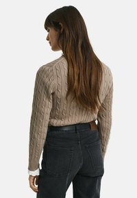 Beige kabelgebreide trui met lange mouwen, van achteren bekeken, in combinatie met donkere spijkerbroek met achterzakken en een leren accent.