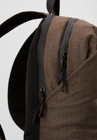 Sac à dos en tissu marron avec une surface texturée, des accents en maille noire et des compartiments à double zip dotés de quincaillerie noire.