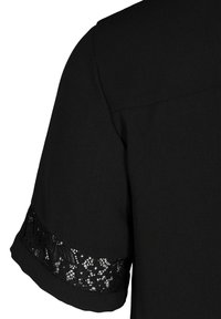 Vêtement noir à manches courtes avec bordure en dentelle près du poignet, montrant la texture du tissu et les détails des coutures sur fond uni.