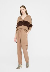 Beige pletený svetr s hnědým chlupatým pruhem, béžové kalhoty s elastickými manžetami a kapsami a vzorované kotníkové boty s blokovými podpatky.