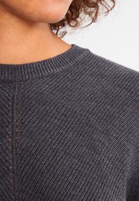 Tummanharmaa neulepaita, jossa on teksturoitu neulos, pyöreä pääntie ja hienovaraiset yksityiskohdat hartioilla. Pehmeä materiaali ribbaiduilla reunoilla.