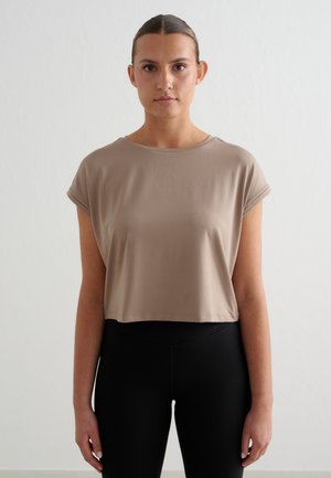 Kobieta z włosami związanymi do tyłu, ubrana w beżową krótki top z krótkim rękawem i czarne legginsy z wysokim stanem, stojąca na tle białego, gładkiego tła.