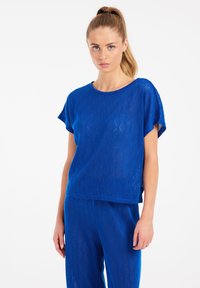 Mėlynas megztinis su deimantų raštu, trumpomis rankovėmis ir laisvu siluetu. Pagamintas iš lengvos medžiagos su tekstūriniu paviršiumi.