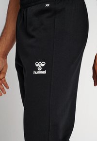 Zwarte joggingsbroek van katoenmix, met een geribbelde tailleband en het witte "hummel" logo nabij de linkerheup. Soepele textuur, casual pasvorm.