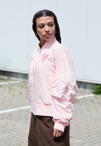 Light pink bomber jacket with a zip front, ribbed cuffs, and embroidered logo. Features gathered sleeves and smooth, shiny fabric. Brown pants.