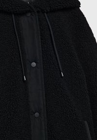 Schwarze Fleece-Jacke mit einer strukturierten Außenschicht, glattem Innenfutter, Druckknöpfen und Kordeln an der Kapuze und dem Saum.