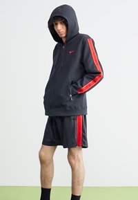 Sweatshirt preto com capuz e riscas laterais vermelhas, bolsos com fecho éclair, combinado com calções pretos correspondentes com detalhes vermelhos. Textura suave.