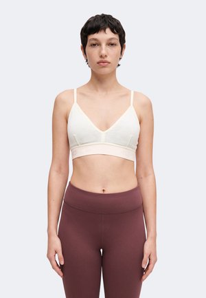 Žena s krátkymi tmavými vlasmi, ktorá má na sebe biely bralette a vysoké bordové legíny, stojaca pred jednoliatym svetlým pozadím.