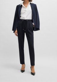 Kvinna iklädd marinblå kavaj hängande över axlarna, vit skjorta med knappar, marinblå byxor med dragsko och svarta spetsiga klackskor framför en enfärgad bakgrund.