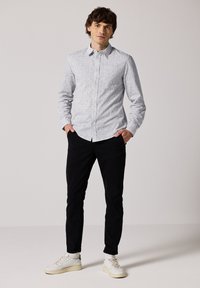 Bílá košile s dlouhými rukávy a jemným černým texturovaným vzorem, spárovaná s černými kalhotami a bílými teniskami. Stojící pozice, ruce v kapsách.