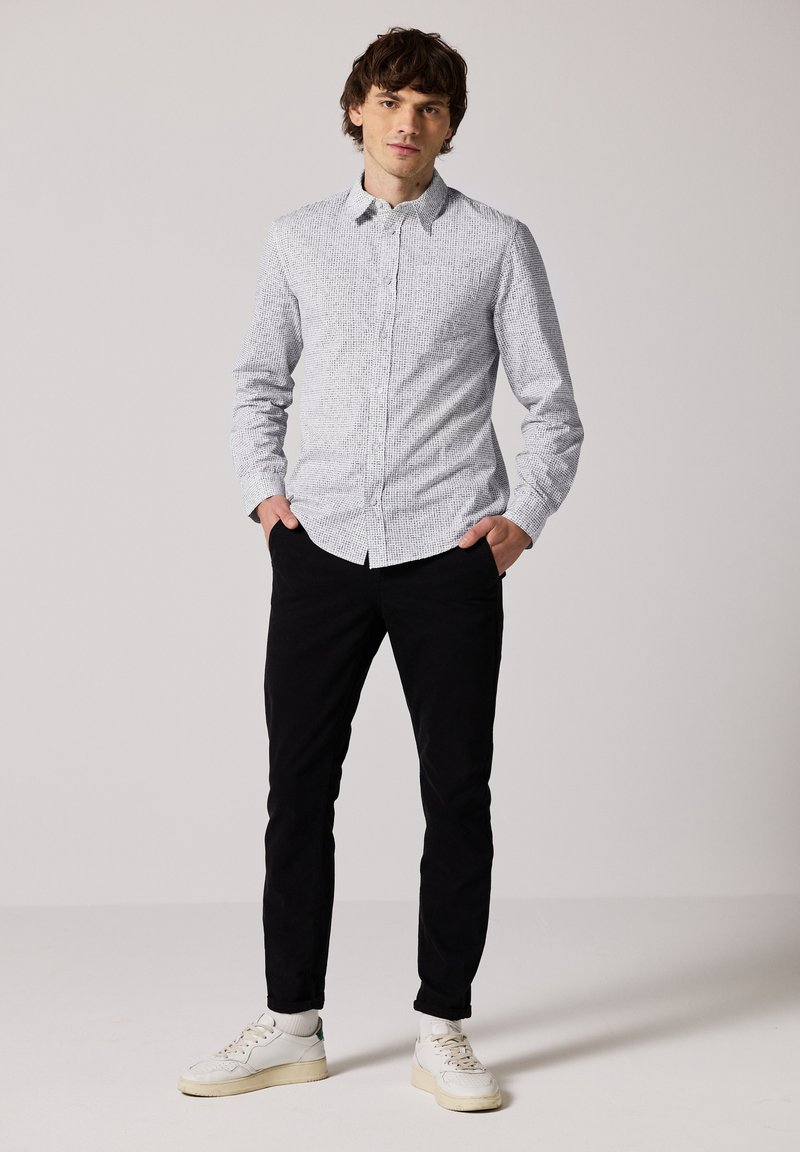 Bílá košile s dlouhými rukávy a jemným černým texturovaným vzorem, spárovaná s černými kalhotami a bílými teniskami. Stojící pozice, ruce v kapsách.