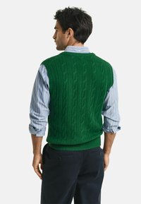 Groene gebreide sweater vest met een textuur van kabelpatroon, ronde halslijn, gedragen over een lichtblauw gestreept shirt, gecombineerd met donkere broek.