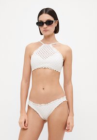 Biely háčkovaný bikiny set s halter krkom, vyznačujúci sa zložitými vzormi a ryhovanými okrajmi, v kombinácii s ladiacimi bikiny spodkami.