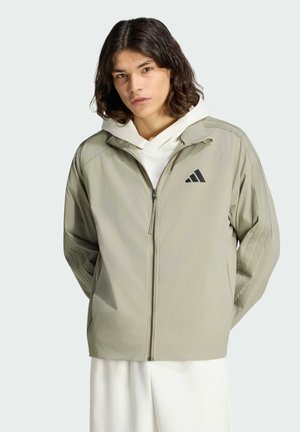 Giovane con capelli castani fino alle spalle che indossa una giacca beige chiaro con zip sopra una felpa bianca con cappuccio, con le mani dietro la schiena.