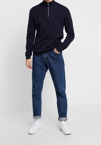 Marineblauwe trui met ritssluiting op de hals en lange mouwen, gecombineerd met blauwe denim jeans. De trui heeft geribbelde manchetten en zoom; de jeans hebben opgerolde mouwen.