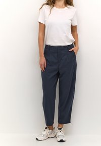 Pantalon plissé bleu marine en tissu léger, doté d'une coupe décontractée et de poches latérales, associé à un t-shirt blanc uni et des baskets blanches.
