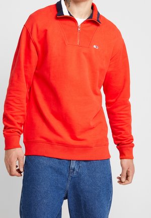 Homme portant un sweat-shirt orange vif à fermeture éclair jusqu’à la poitrine avec un petit logo sur la poitrine, associé à un jean denim bleu, tatouage visible sur le cou.