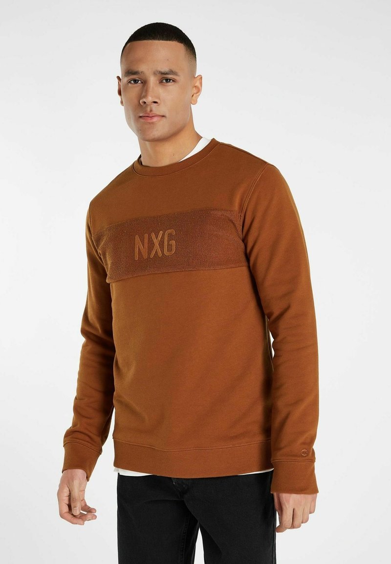 Brun sweatshirt med en texturerad horisontell rand med "NXG"-logotyp. Långa ärmar, rund halsringning, ribbade muddar och fåll. Svarta jeans.