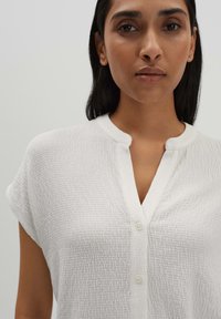 Camicia bianca con bottoni, in tessuto texture, maniche corte e colletto alla mandarino. Presenta bottoni beige e un tessuto leggero.