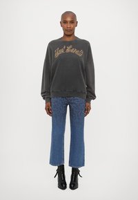 Γκρι oversized φούτερ με τη φράση "Just Casuals" σε χρυσή γραφή, συνδυασμένο με φαρδιά μπλε τζιν και μαύρες μπότες μέχρι τον αστράγαλο. Λείες υφασματικές υφές.