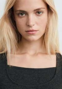 Haut noir ajusté avec un col carré et une texture lisse, porté par un mannequin aux cheveux clairs devant un fond uni.