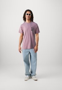 Camiseta de manga corta de color púrpura claro con estampado gráfico en el lado izquierdo del pecho, combinada con jeans rectos de color azul claro y zapatillas blancas.