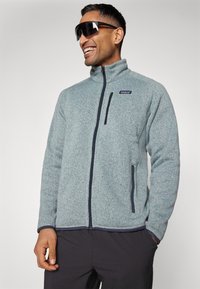 Casaco de fleece cinza claro com gola alta, zipper frontal, detalhes em azul-marinho e bolsos laterais. Tecido texturizado com um ajuste casual.