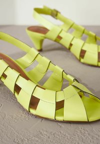 Keltaiset nahkaiset slingback-kengät, joissa on ristikkäiset hihnat, suljettu kärki ja kapeneva korko teksturoidulla beigealla pinnalla.