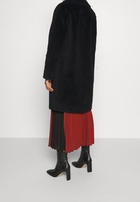 Svart kappa med en strukturerad yta och bred krage, kombinerad med en plisserad kjol i svart och röd, samt svarta klackstövlar.