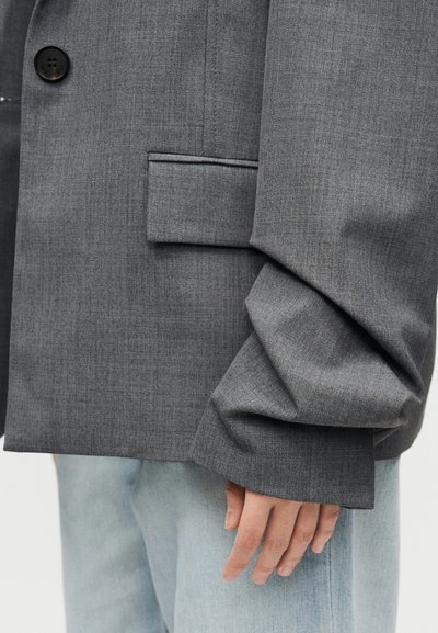 Άτομο που φοράει υπερμεγέθη γκρι blazer με πέπλα μανίκια και ανοιχτό μπλε τζιν, χέρι χαλαρό στο πλάι.